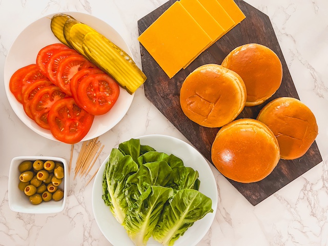 Ingredients for Halloween monster burgers including brioche buns, burger patties, provolone cheese, pickles, lettuce, tomatoes, and green olives.
