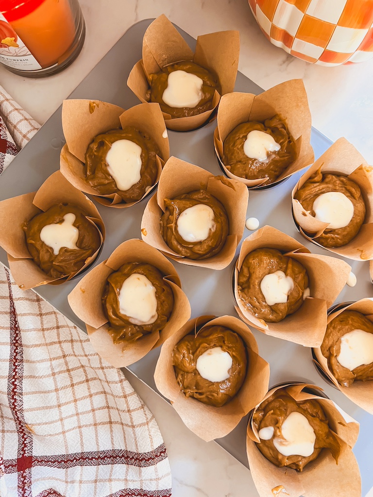 Cream cheese filling being piped into the center of pumpkin muffin batter in muffin cups.