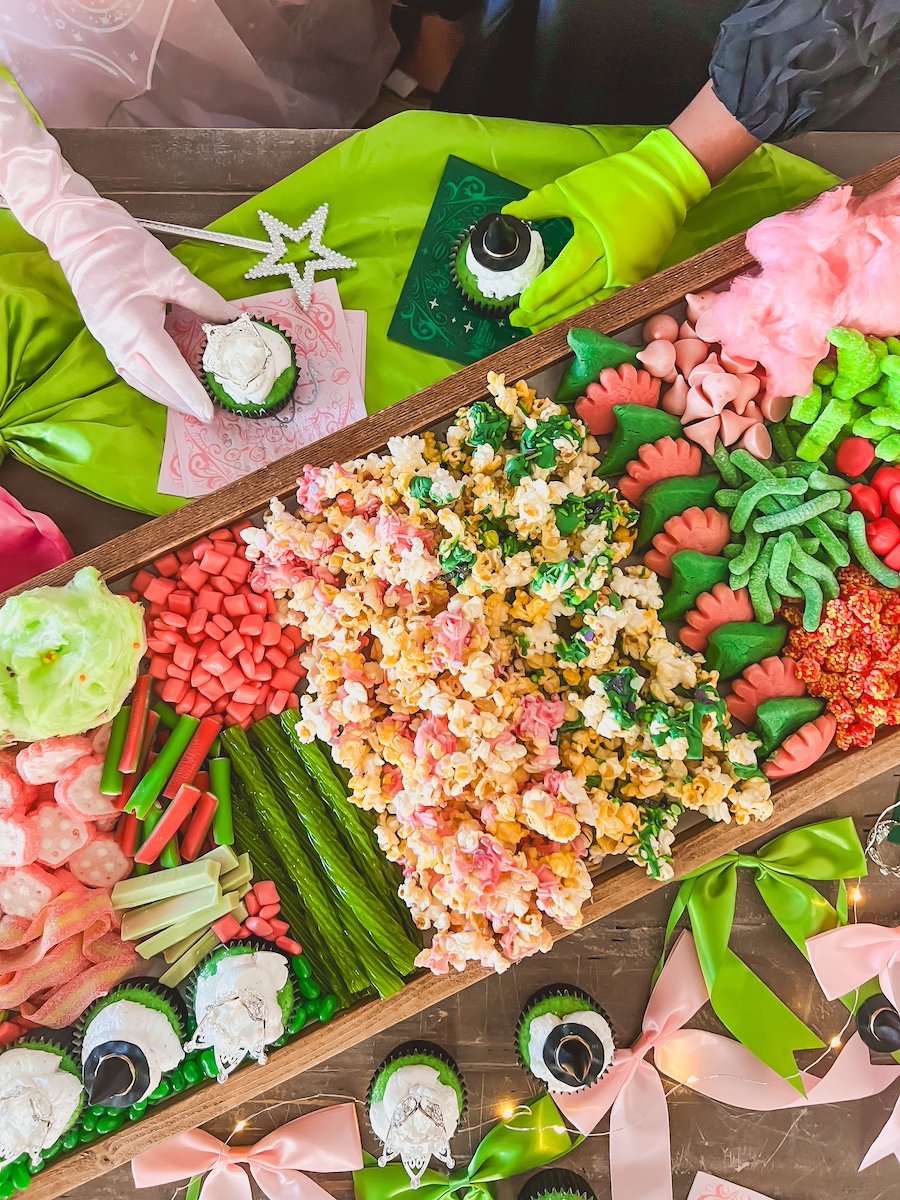 bird eye view of wicked movie night board with a pink glove hand and a green alfaba hand grabbing a cupcake