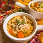 Close up image of a white crock with slow cooker chicken noodle soup and another crock in the background.