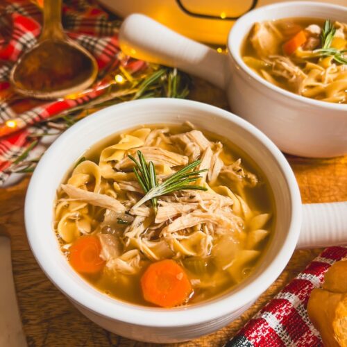 Close up image of a white crock with slow cooker chicken noodle soup and another crock in the background.