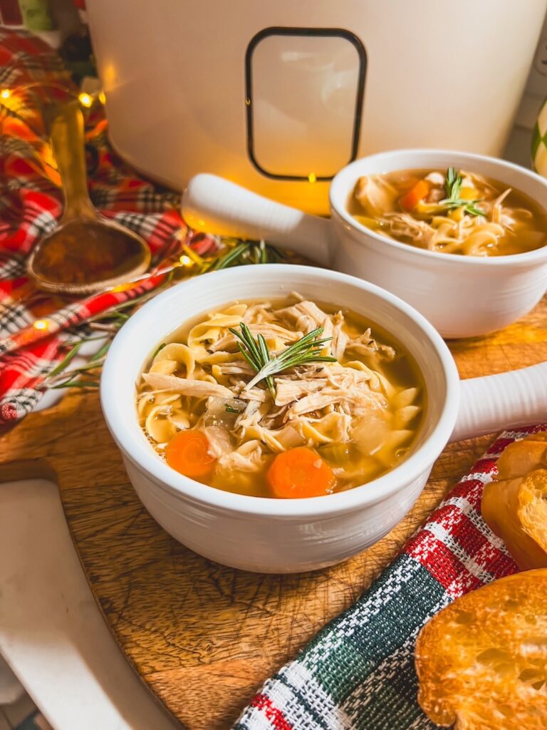 Two white crocks of slow cooker chicken noodle soup with the crockpot in the background.