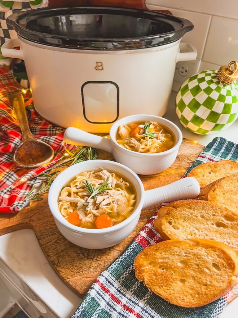 Two white crocks of slow cooker chicken noodle soup with the crockpot in the background.