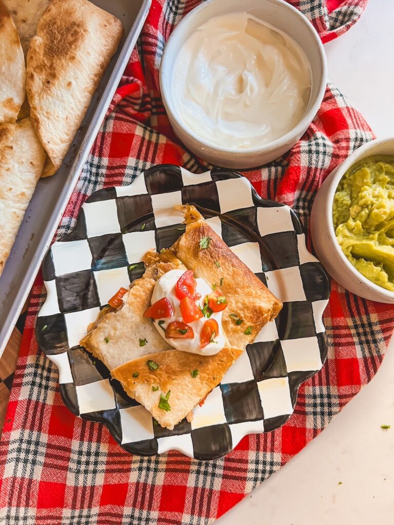 Overhead image of a slice of sheet pan chicken fajita quesadilla on a checkered plate with sour cream and guacamole. 