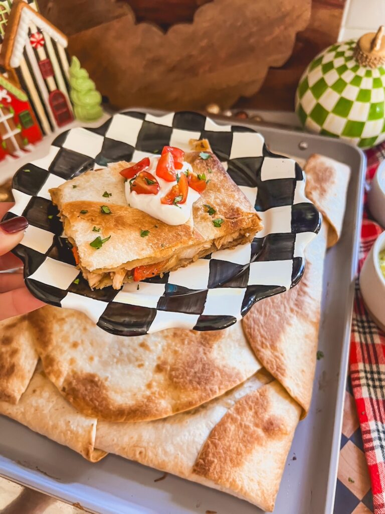 A slice of chicken fajita quesadilla on a checkered plate with the chicken fajita sheet pan quesadilla in the background. 