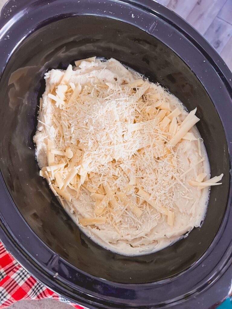 Shredded cheese, butter, and cream being added to mashed potatoes in the slow cooker. 