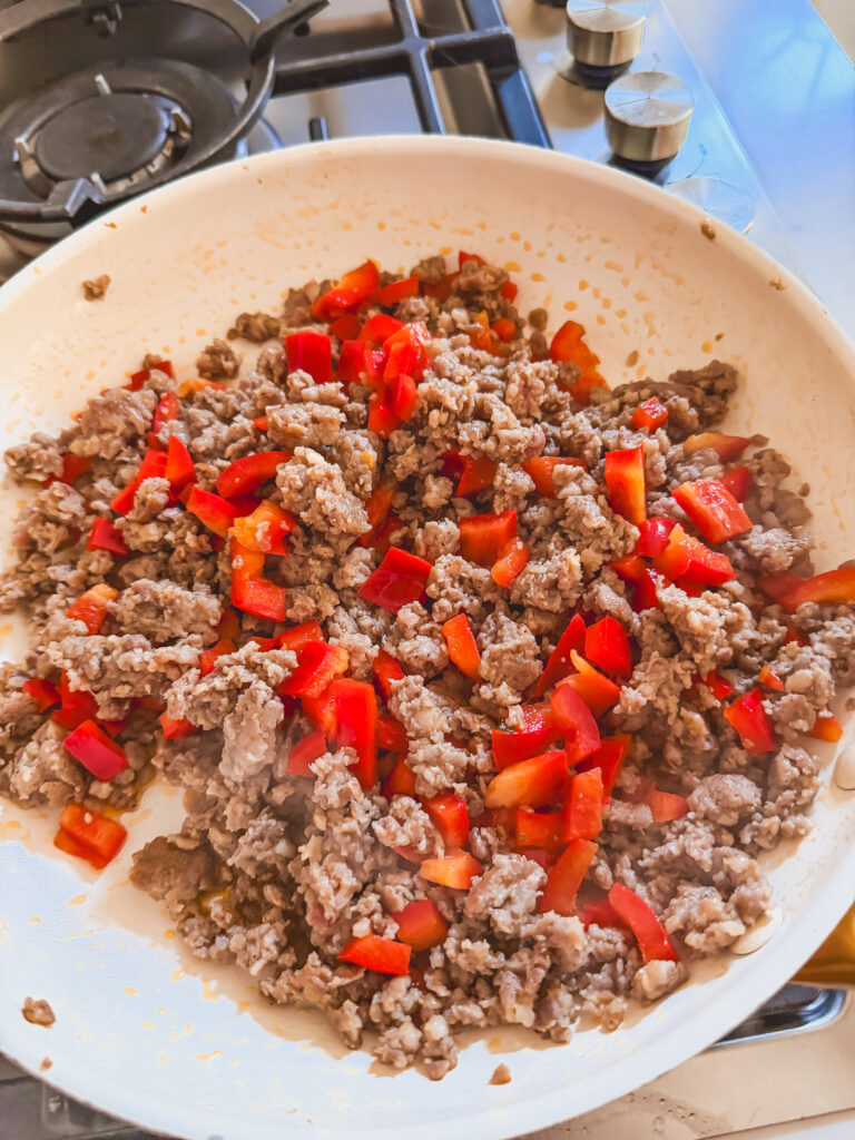 A white skillet with breakfast sausage and red bell pepper cooking on the stovetop.