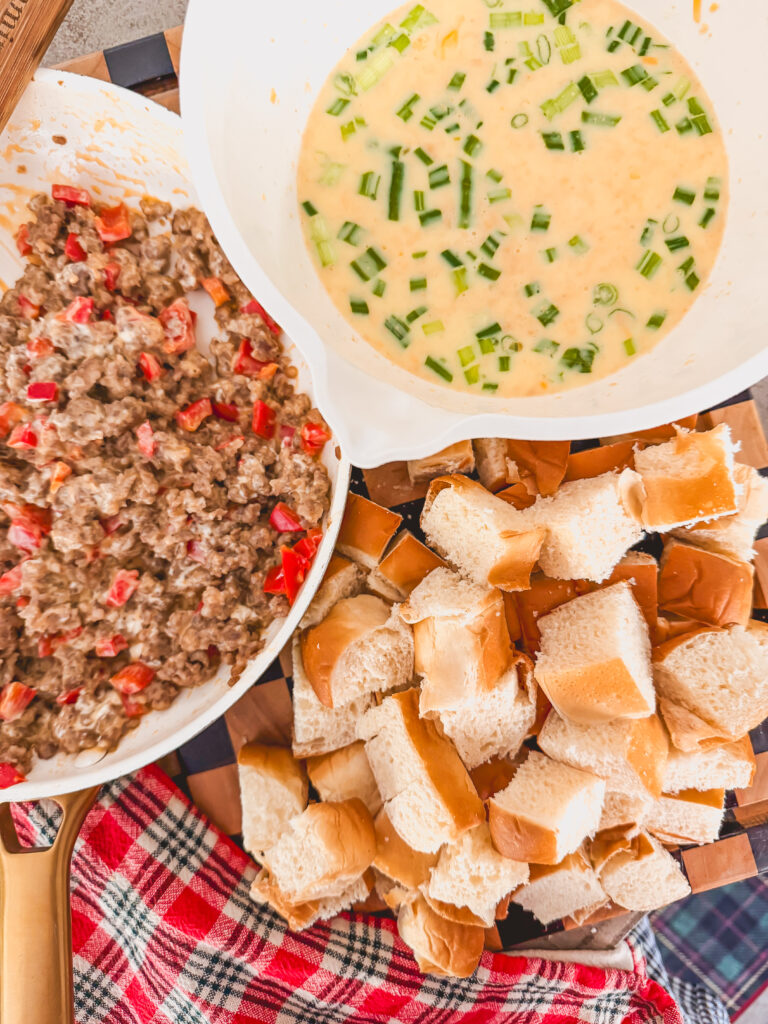 sausage mixture in a white skillet, with egg mixture in a white bowl, and Kings Hawaiian rolls cut into cubes on a plaid linen.