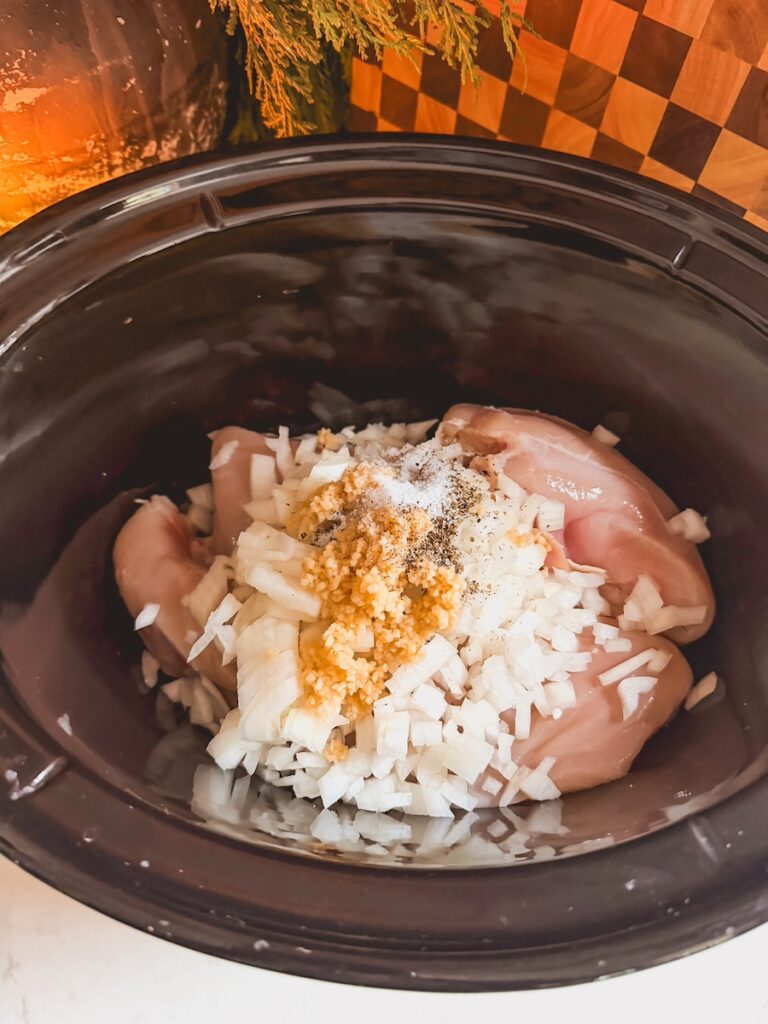A crockpot with raw chicken, garlic, spices, and onions.