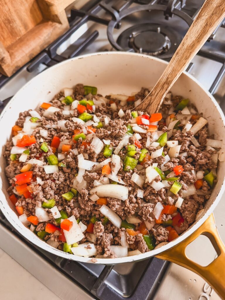 A white saute pan cooking ground beef, peppers, and onions with a wooden spoon on the stovetop.