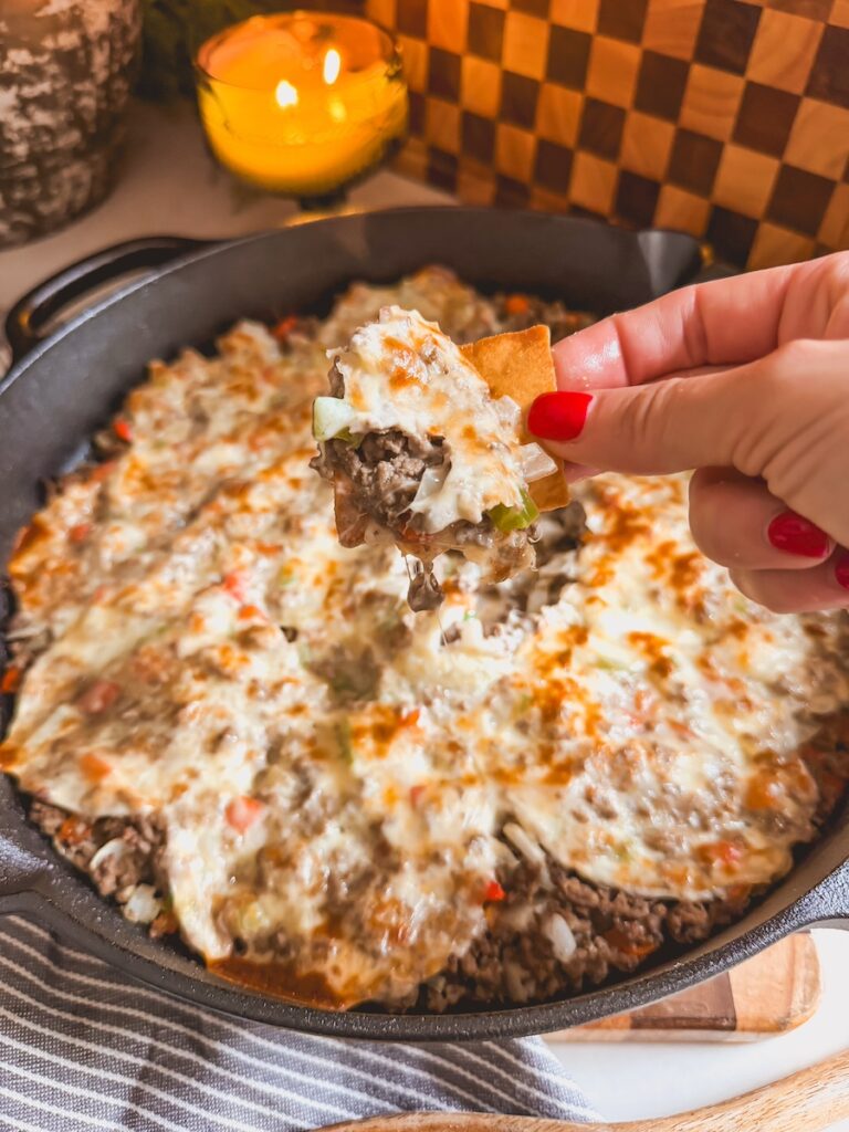 A hand scooping out philly cheesesteak dip from a cast iron skillet.