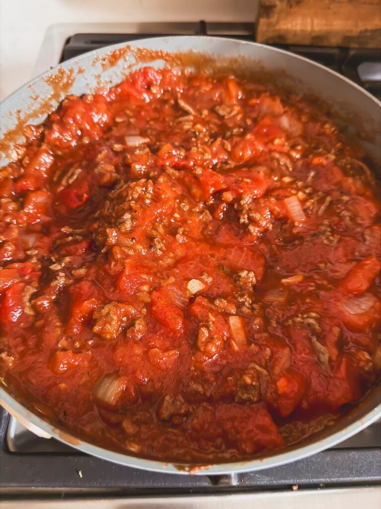 A skillet with ground beef, tomatoes, and onion cooking on the stovetop.