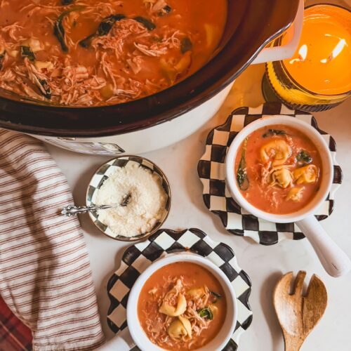 A slow cooker of chicken tortellini soup with two white bowls of soup on black and white checkered plates.