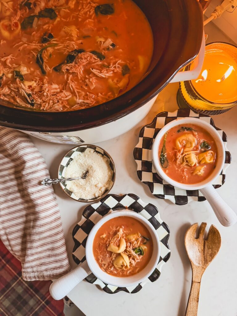 A slow cooker of chicken tortellini soup with two white bowls of soup on black and white checkered plates.