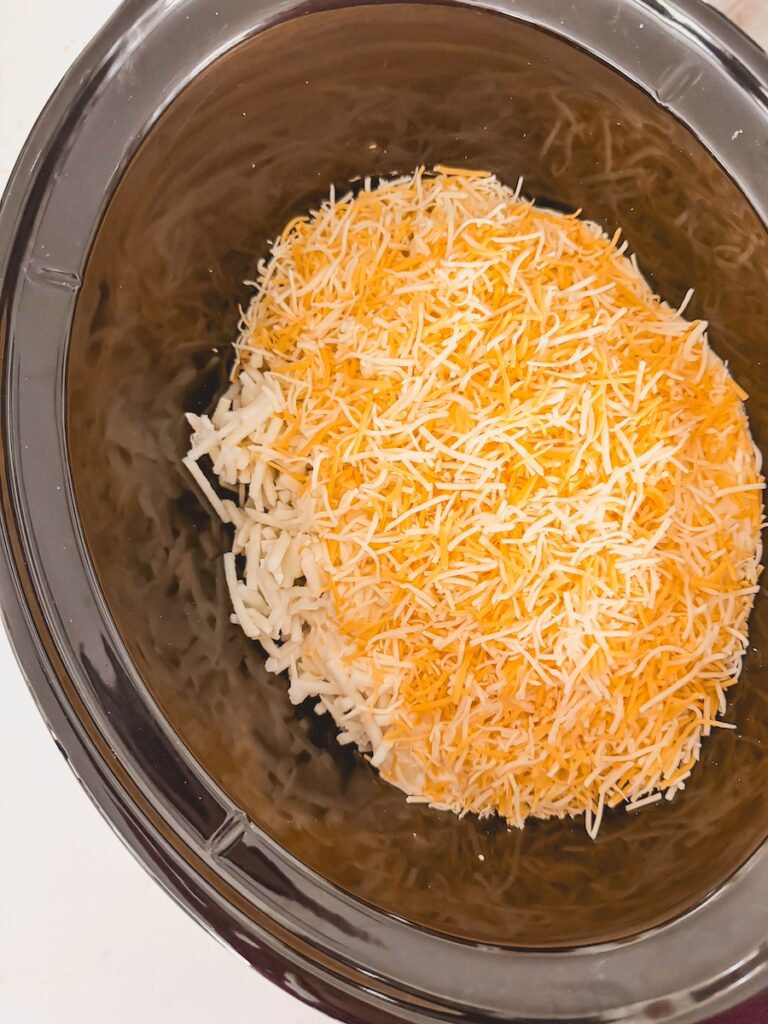 uncooked frozen hashbrowns with creamy sauce and grated cheese on top in bottom of slow cooker