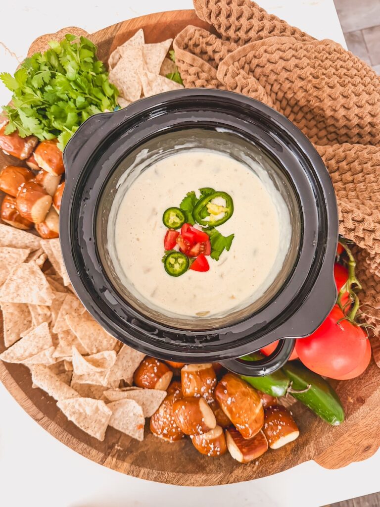 overhead view of cooked crock pot white queso in slow cooker on board with chips, pretzels and vegetables around it