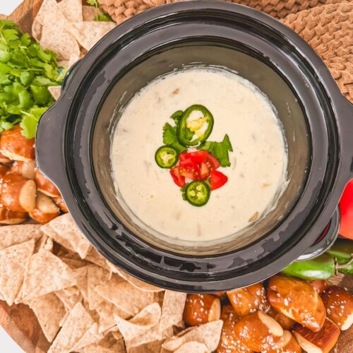 overhead view of cooked crock pot queso in slow cooker in center of board with chips, pretzels, vegetables and dish towel around it
