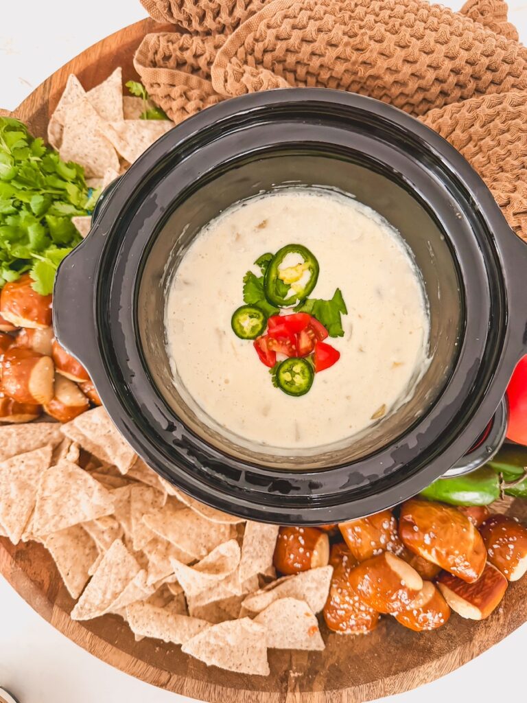 overhead view of cooked crock pot queso in slow cooker in center of board with chips, pretzels, vegetables and dish towel around it