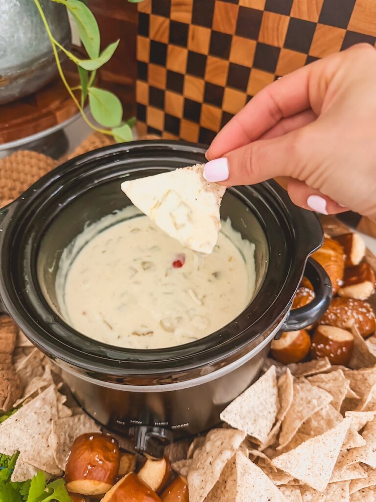 right hand holding a chip that has been dipped into crock pot queso with chips and pretzels in background