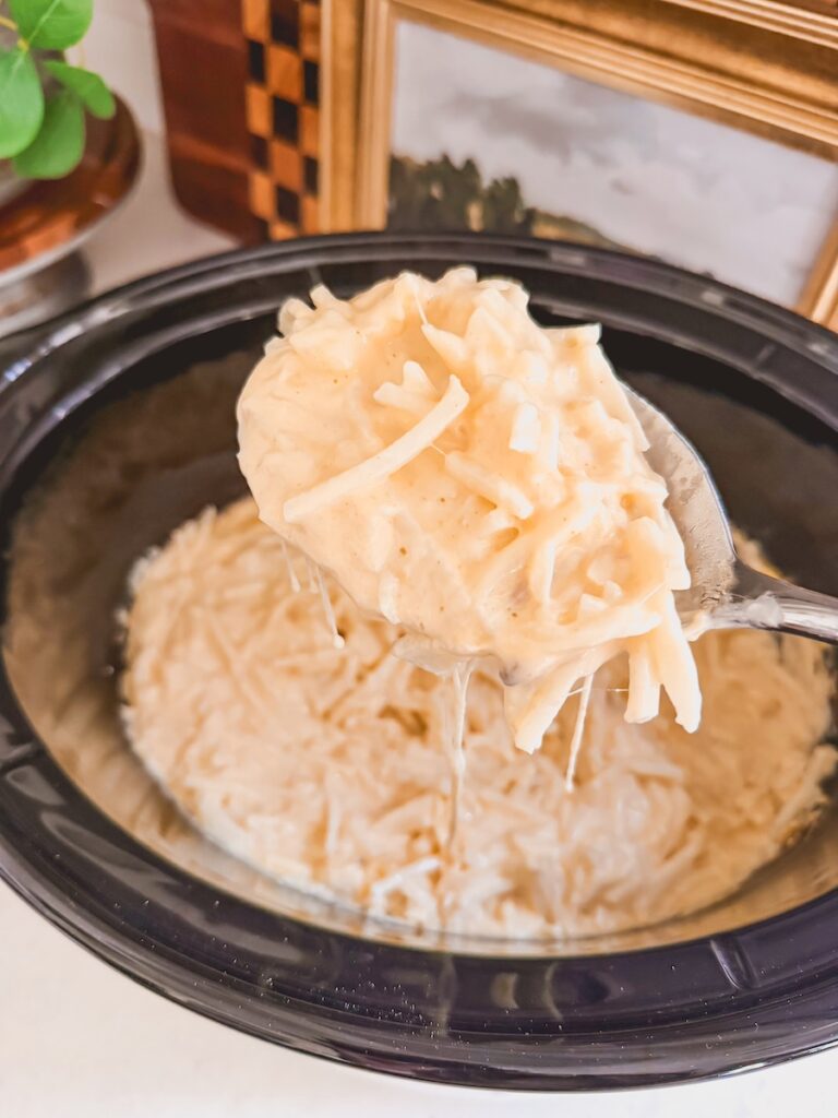a spoonful of cooked cheesy potatoes coming out of slow cooker with cheese pull, with slow cooker in background