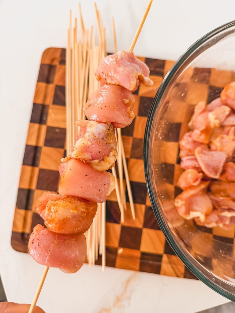 Chicken teriyaki pieces threaded onto a wooden skewer. A glass bowl with additional chicken pieces is in the background.
