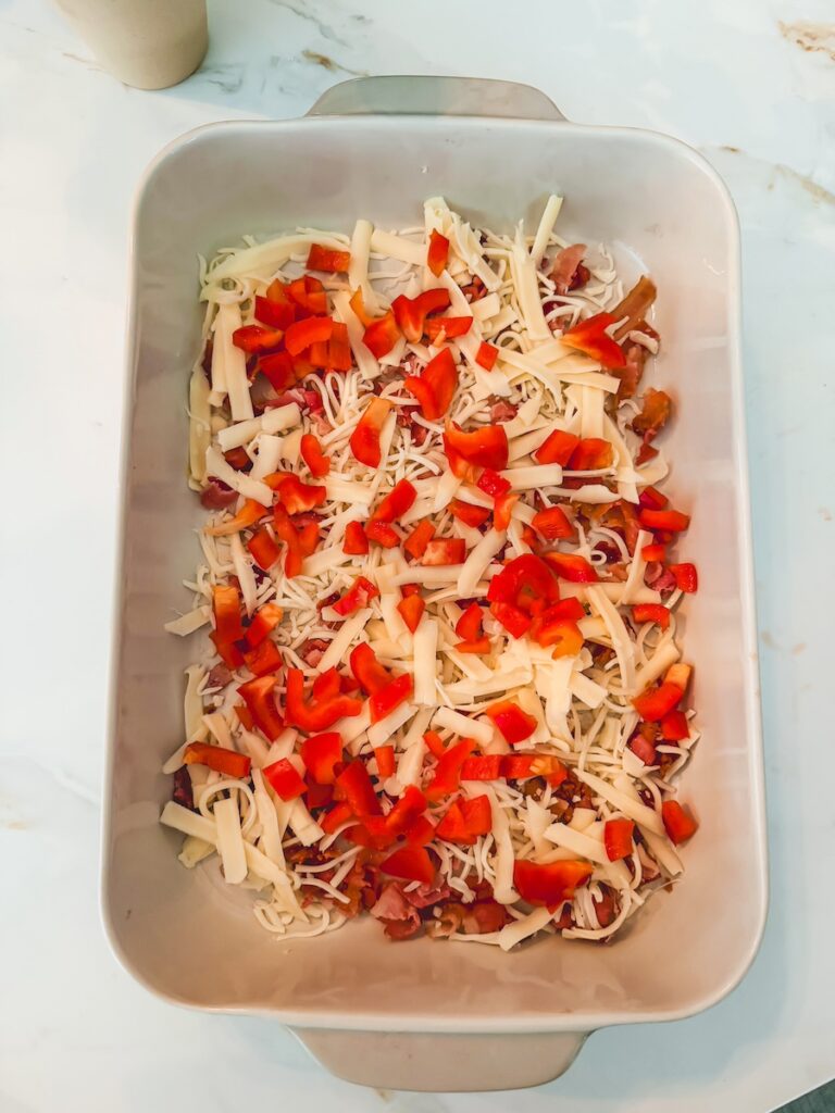 A white baking dish with bacon, shredded cheese, and bell pepper.