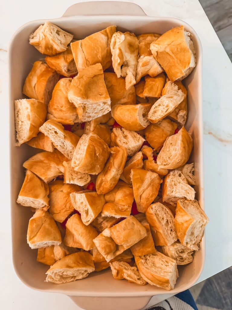 A white baking dish with cut up croissants, shredded cheese, and bell pepper for croissant breakfast casserole.