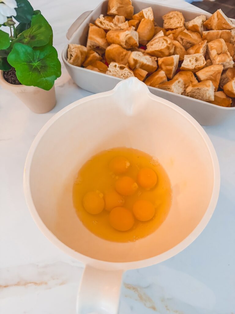A white bowl with eggs and a baking dish with cut up croissants for croissant breakfast casserole.