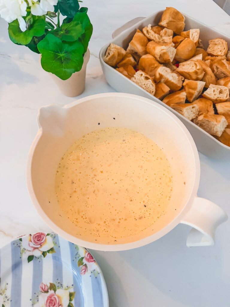 A white bowl with whisked eggs with a white baking dish with cut up croissants in the background.