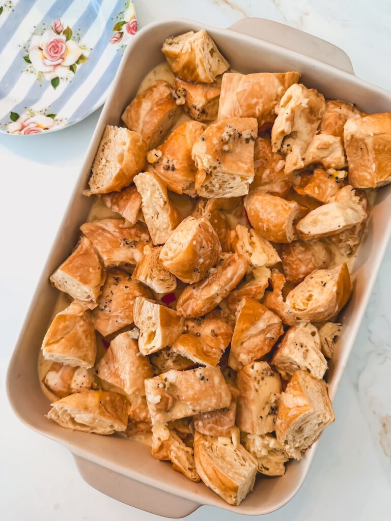 A white baking dish with croissant breakfast casserole before baking and a striped blue plate in the background.