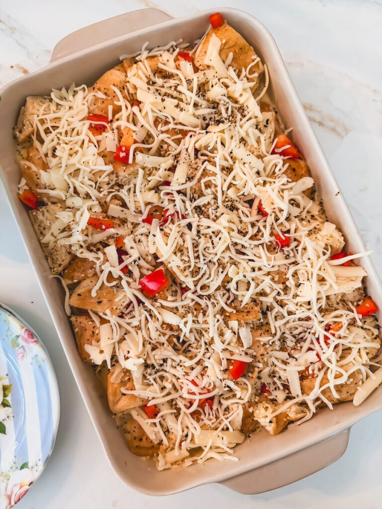 A white baking dish with raw croissant breakfast casserole with shredded cheese on top before baking.