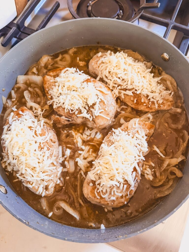 chicken breasts topped with cheese in a pan with caramelized onions and broth