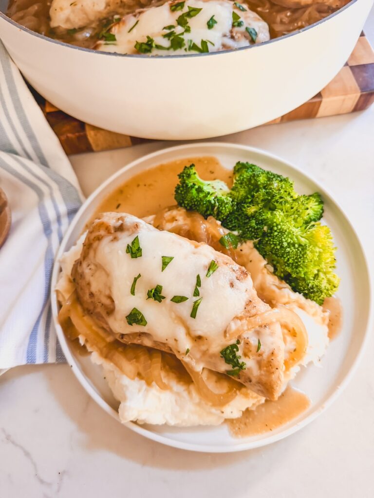 plated serving of french onion chicken with potatoes and broccoli. Pot of french onion chicken on cutting board in background.