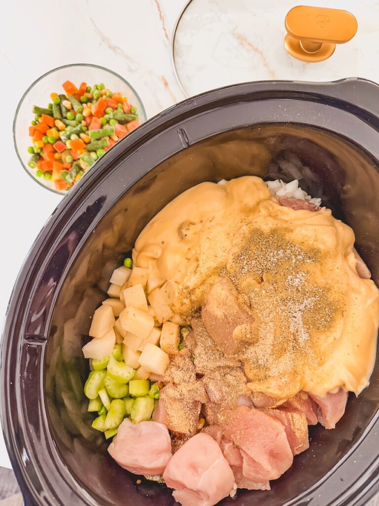 slow cooker chicken pot pie ingredients uncooked, in slow cooker. frozen veggies in a glass bowl and lid in background.
