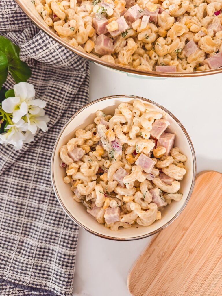 Ham pasta salad in large bowl with small individual serving in smaller bowl next to it. Cutting board and checked napkin in background.