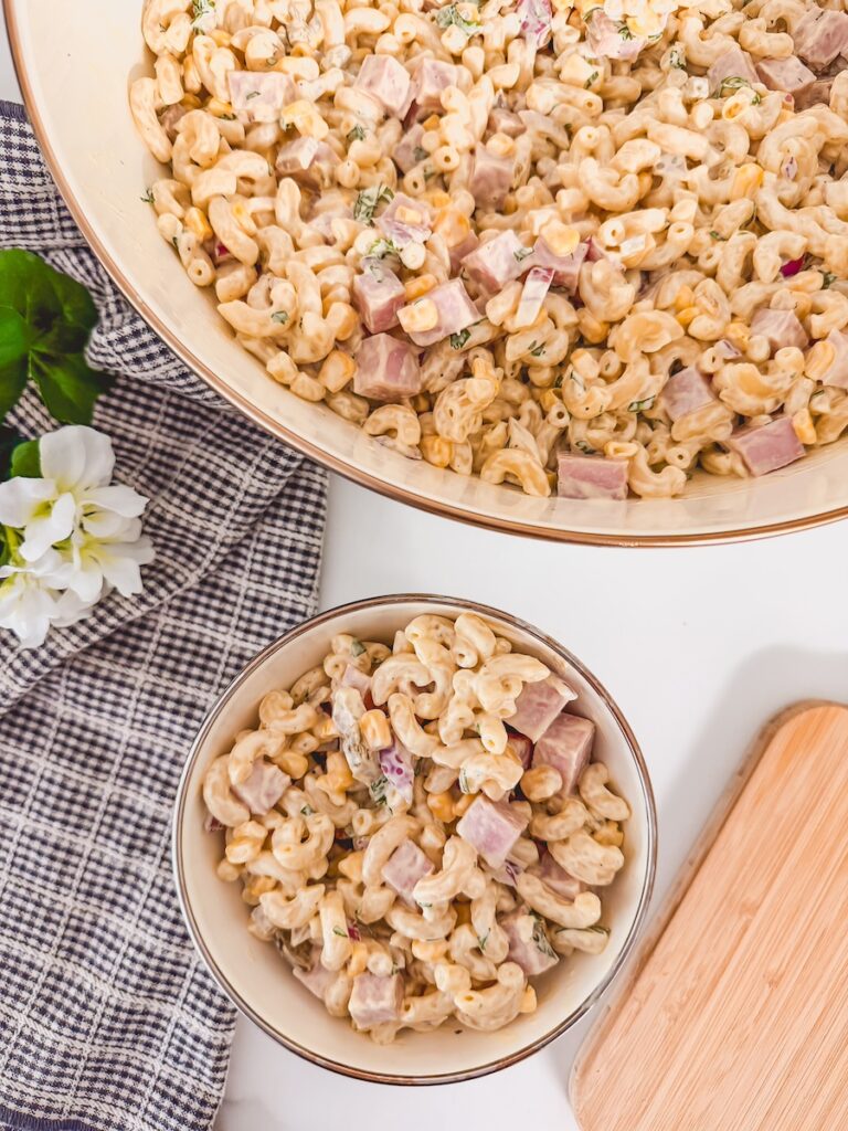 Ham pasta salad in large bowl with small individual serving in smaller bowl next to it. Cutting board and checked napkin in background
