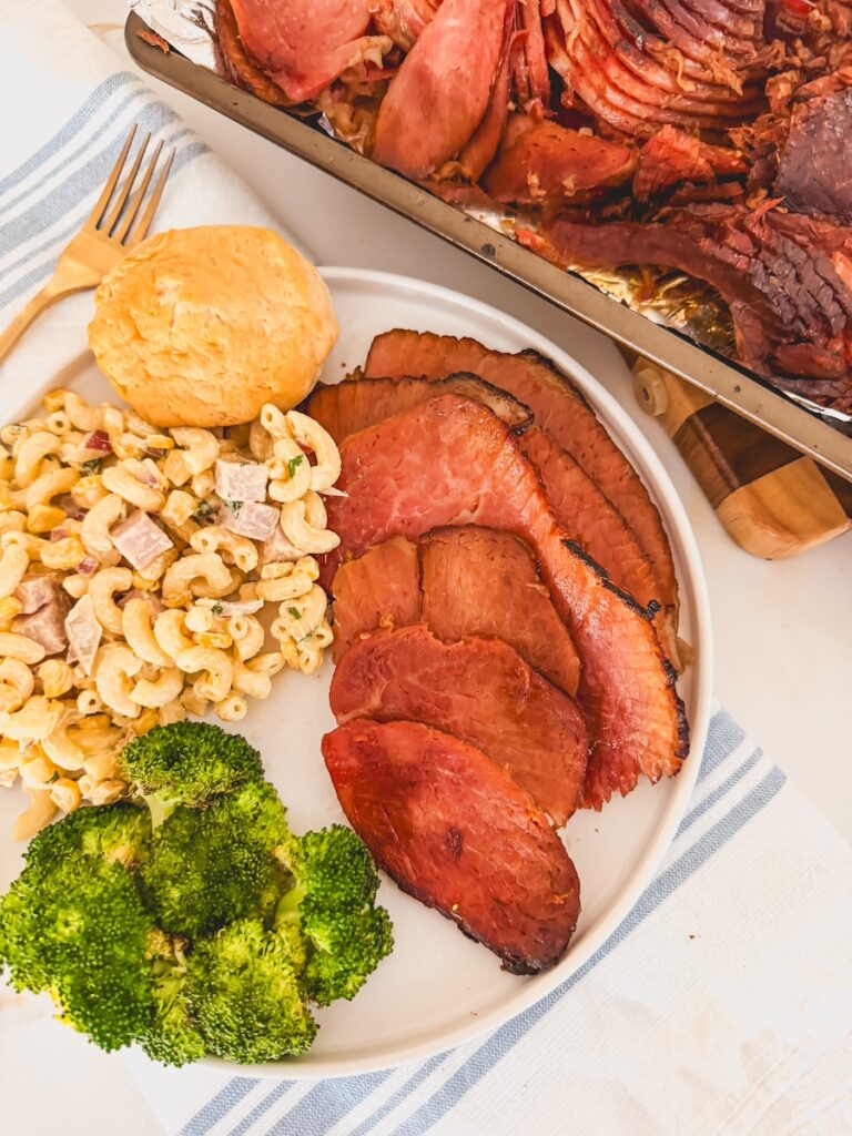 cola ham plated on white plate with broccoli and pasta salad. baking sheet with ham on it next to plate