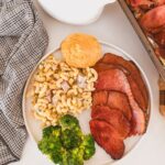 serving of ham on plate with broccoli and pasta salad, baking sheet of sliced ham and saucer of glaze