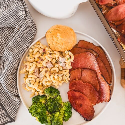 serving of ham on plate with broccoli and pasta salad, baking sheet of sliced ham and saucer of glaze