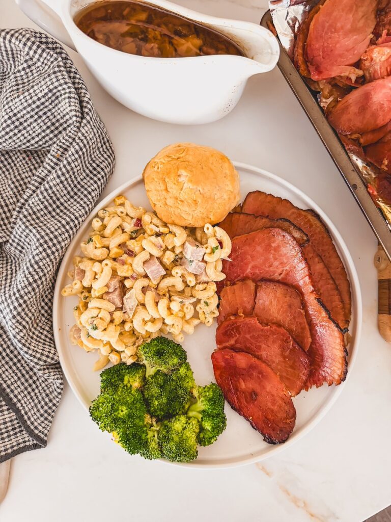 serving of ham on plate with broccoli and pasta salad, baking sheet of sliced ham and saucer of glaze