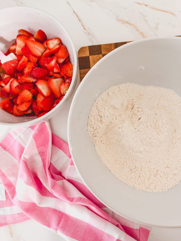 sliced strawberries in white bowl next to flour and dry ingredients in larger white bowl. striped pink towel and cutting board in background.