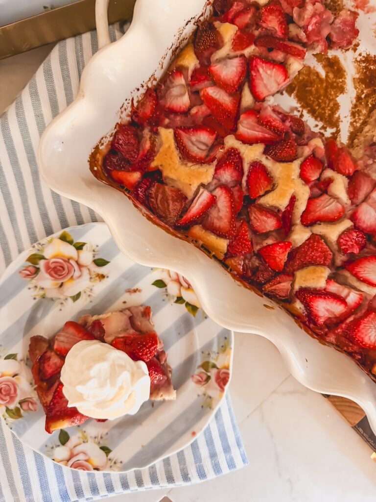 baked strawberry cobbler in white baking dish with a serving scooped out and plated on a striped and floral plate next to it with whipped topping