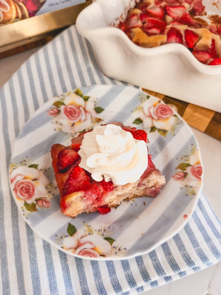 plated serving of strawberry cobbler topped with whipped cream. strawberry cobbler in baking dish in background.