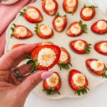 deviled strawberries with cheesecake filling on a tray with a hand holding one