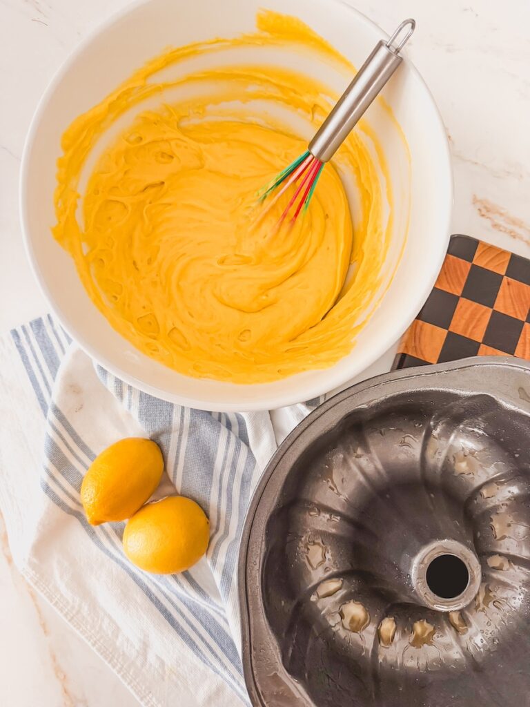 cake batter in bowl with whisk next to greased bundt pan. lemons on towel next to bowl and pan.