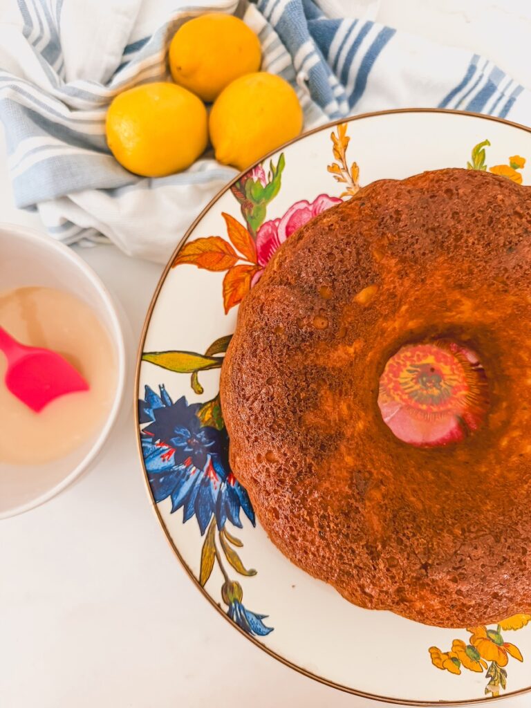 baked and unglazed lemon bundt cake with glaze in a bowl next to it and lemons in the background