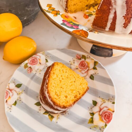 closer shot of slice of plated lemon bundt cake on flower plate next to whole bundt cake with slice cut out. Lemons on table in background.