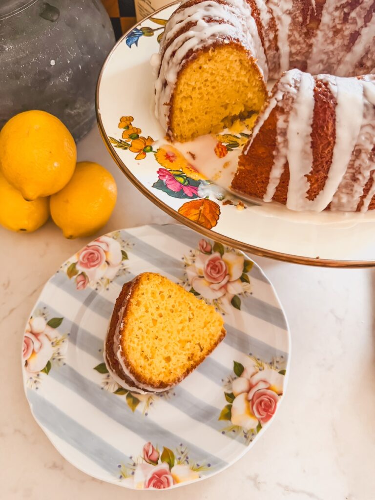 slice of plated lemon bundt cake on flower plate next to whole bundt cake with slice cut out. Lemons on table in background.