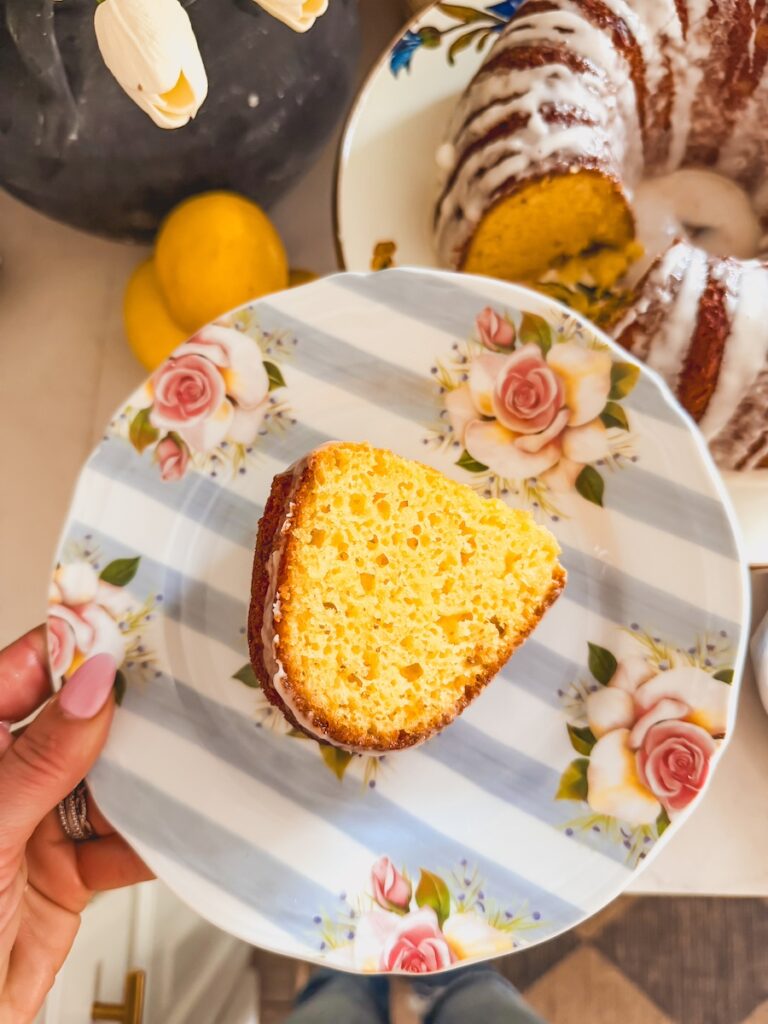 slice of lemon bundt cake on flower plate with hand holding plate. bundt cake in background.