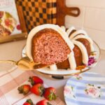 slice of strawberry bundt cake on gold cake server (side profile). bundt cake on cake stand and fresh strawberries in background.